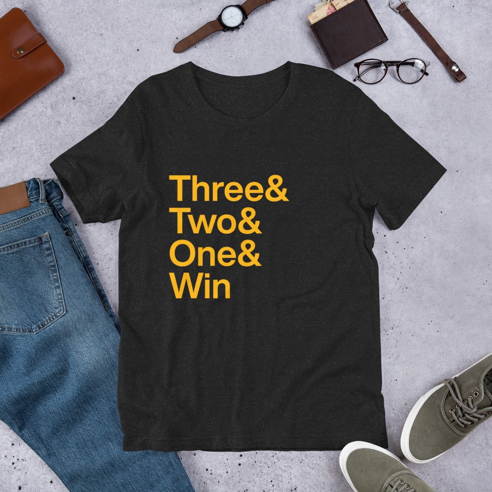 black t-shirt with gold lettering reading Three and Two and One and Win on the ground amid other clothing items