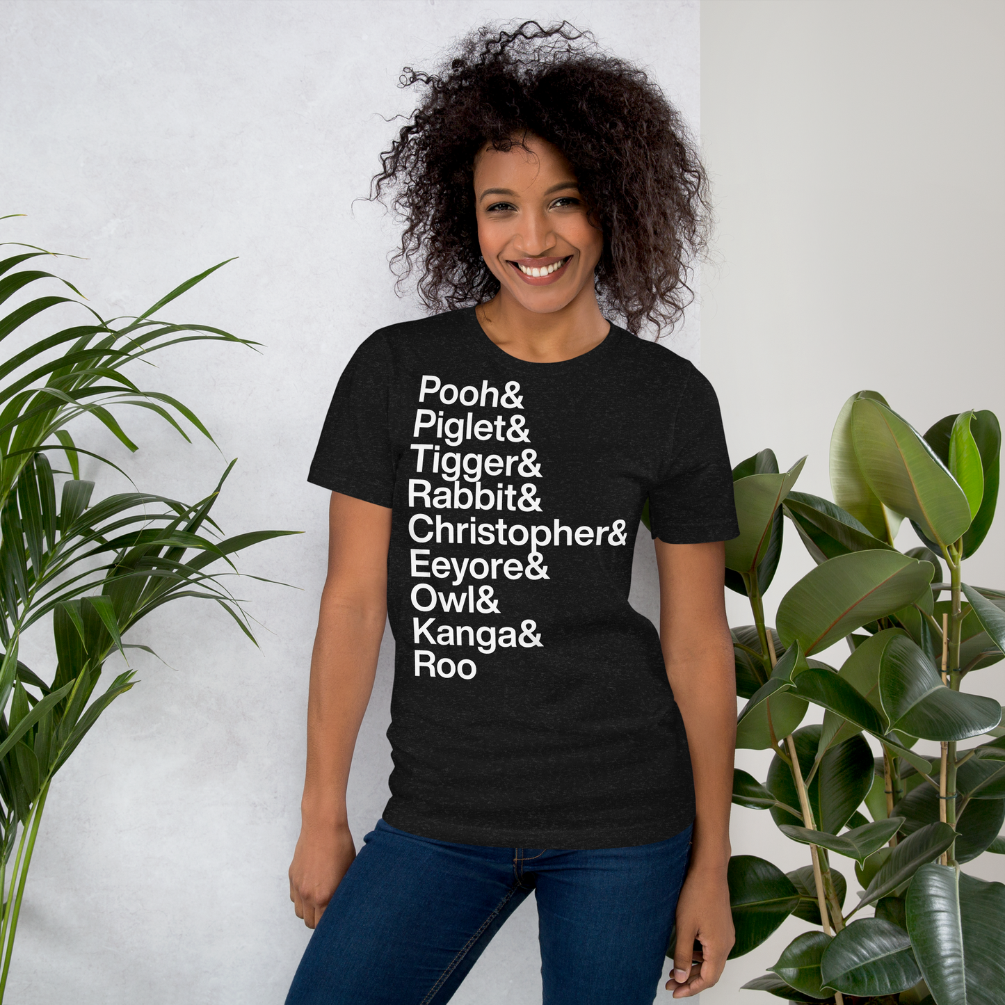 Young woman wearing black t-shirt with white lettering reading Pooh and Piglet and Tigger and Rabbit and Christopher and Eeyore and Owl and Kanga and Roo standing between two plants