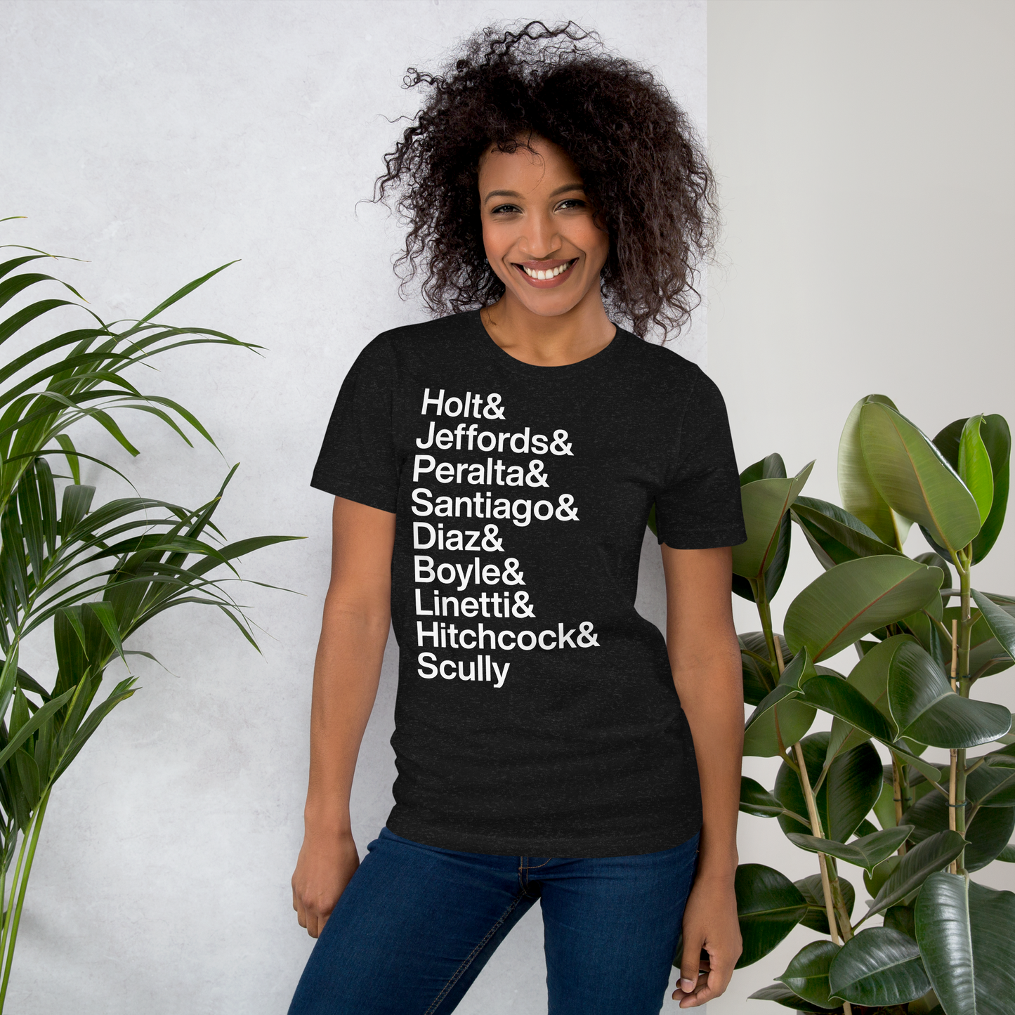 Young woman wearing a black t-shirt with white lettering reading Holt and Jeffords and Peralta and Santiago and Diaz and Boyle and Linetti and Hitchcock and Scully standing next to a plant