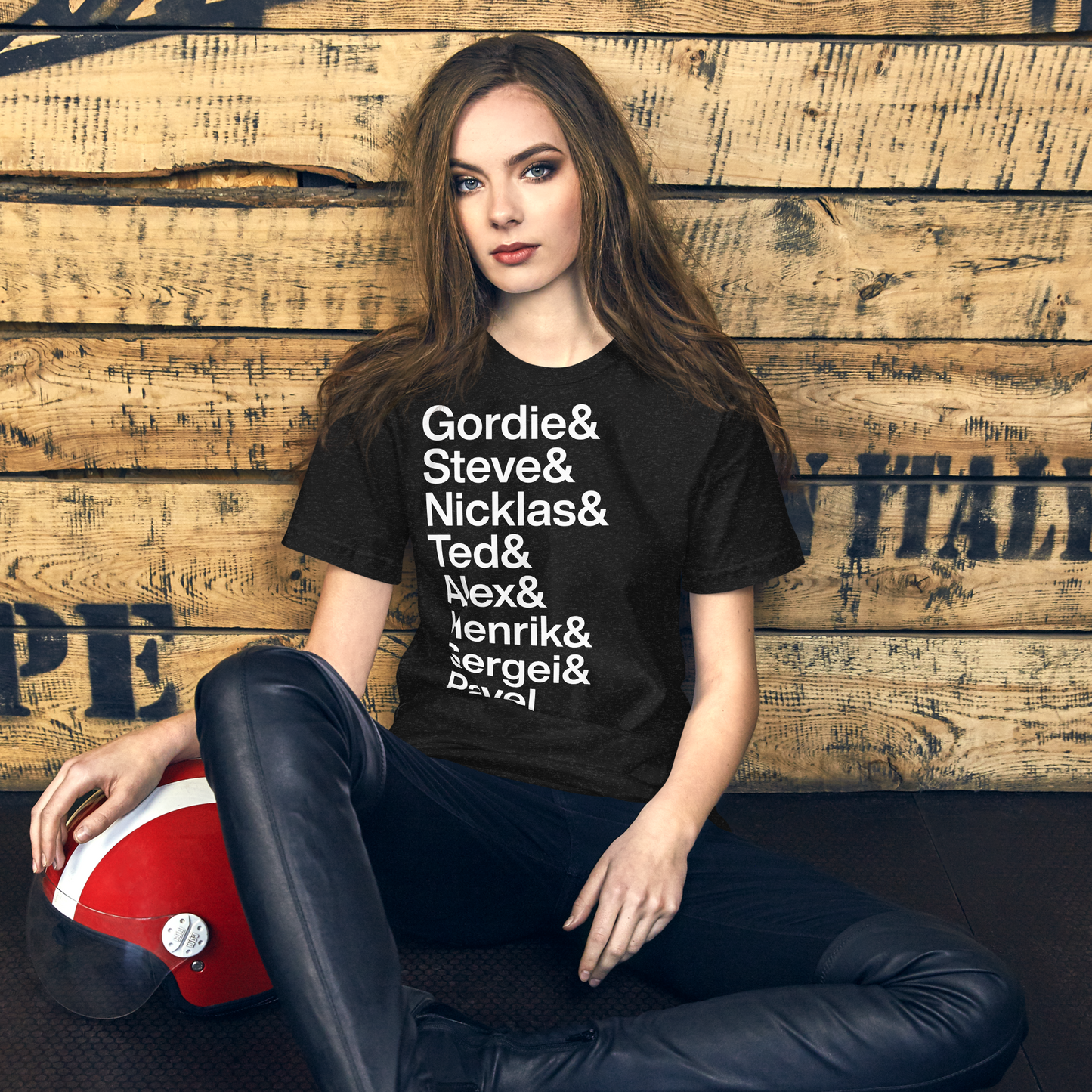Young woman sitting against wall wearing a black t-shirt with white lettering listing GOATs of Red Wings