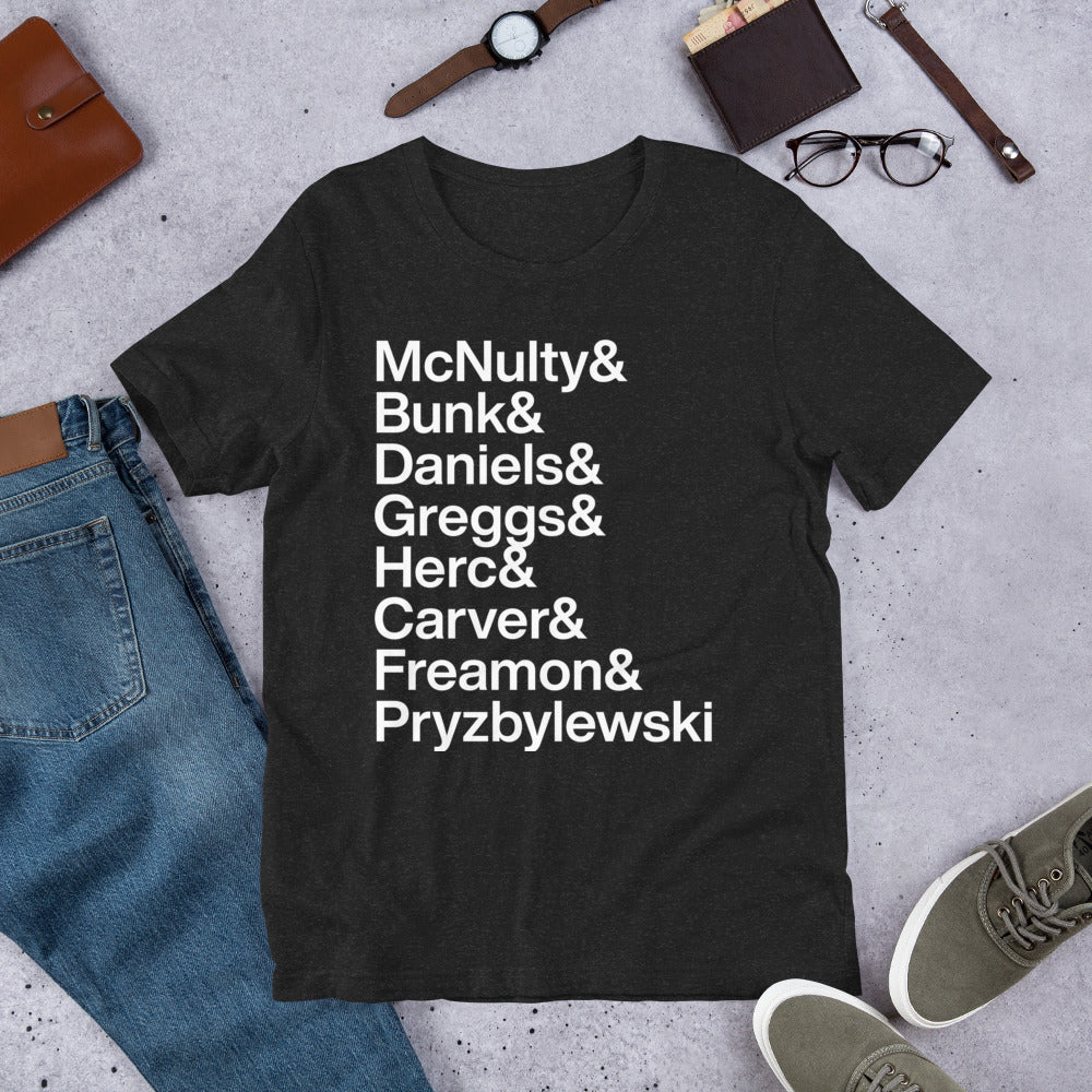 black t-shirt with white lettering reading McNulty and Bunk and Daniels and Greggs and Herc and Carver and Freamon and Pryzbylewski on ground with other apparel