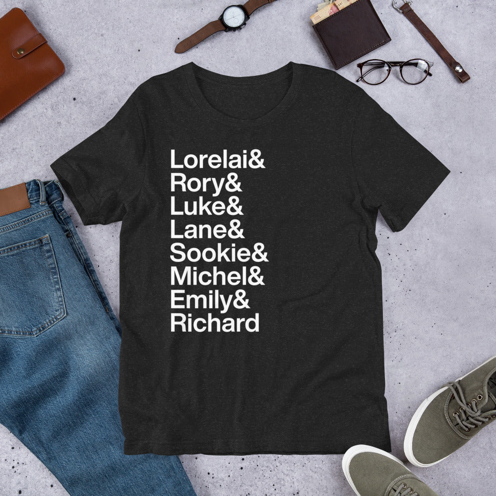 Black t-shirt with white text reading Lorelai and Rory and Luke and Lane and Sookie and Michel and Emily and Richard; t-shirt is resting on ground amidst other clothing