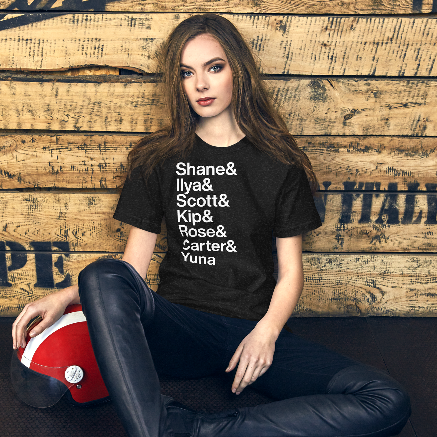 Woman wearing a black t-shirt with names printed on it, sitting against a wooden wall.