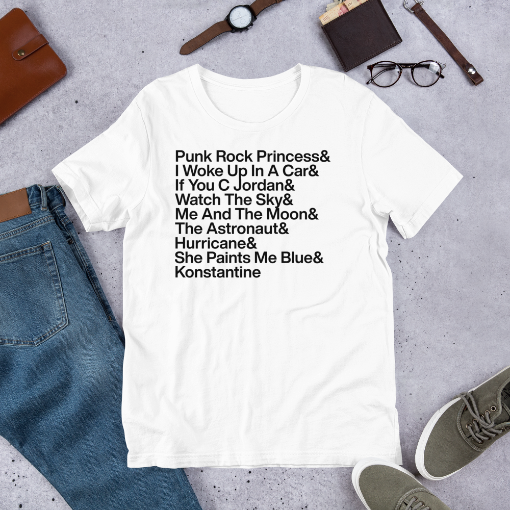 White t-shirt with text on a gray surface with jeans, shoes, and accessories.